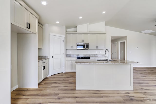 kitchen with a sink, light wood finished floors, stainless steel microwave, lofted ceiling, and a center island with sink