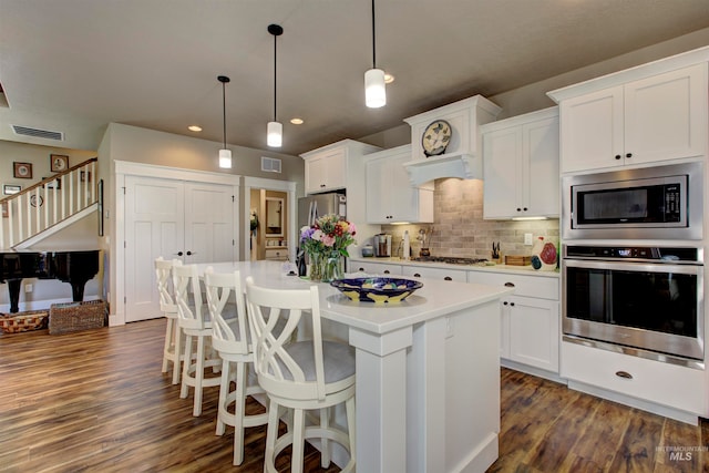 kitchen featuring white cabinets, an island with sink, appliances with stainless steel finishes, dark wood-type flooring, and decorative light fixtures