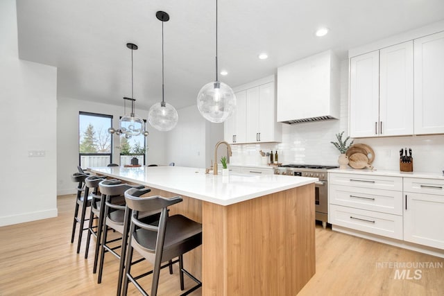 kitchen with wall chimney range hood, high end range, pendant lighting, a kitchen island with sink, and white cabinets