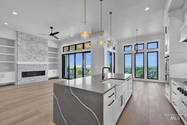 kitchen featuring dark stone countertops, built in features, an island with sink, a stone fireplace, and white cabinets