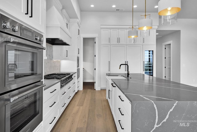 kitchen with white cabinets, appliances with stainless steel finishes, decorative light fixtures, dark stone countertops, and premium range hood