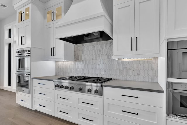 kitchen featuring white cabinets, appliances with stainless steel finishes, light hardwood / wood-style floors, backsplash, and custom range hood