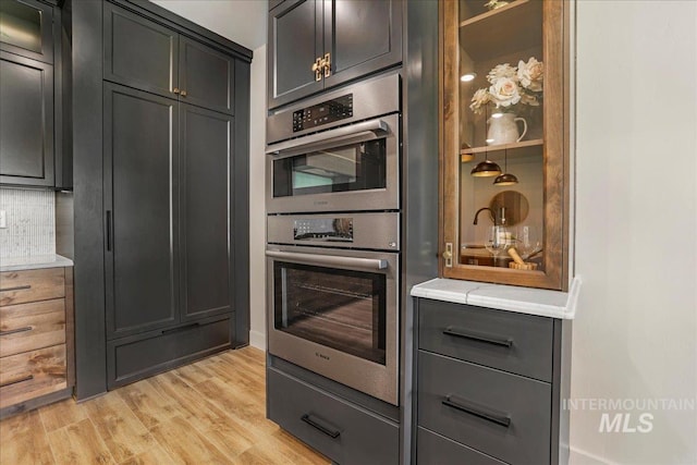 3160 Crescent Moon Rd, Twin Falls, ID 83301 - kitchen with stainless steel double oven, light wood finished floors, tasteful backsplash, light countertops, and glass insert cabinets