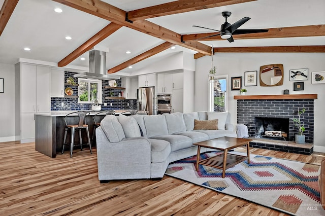 living room with light wood finished floors, ceiling fan, vaulted ceiling with beams, and a brick fireplace