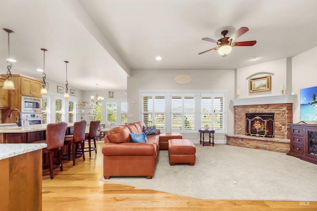 living room featuring light wood-type flooring, ceiling fan with notable chandelier, a wealth of natural light, and a stone fireplace