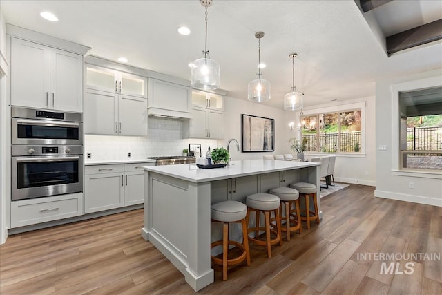 kitchen with decorative light fixtures, tasteful backsplash, light countertops, glass insert cabinets, and an island with sink