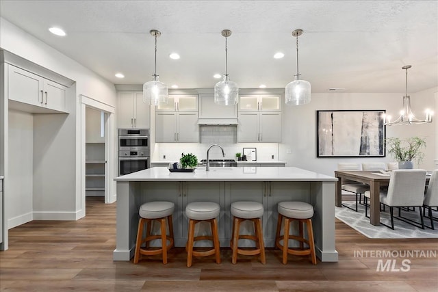 kitchen featuring double oven, a kitchen island with sink, pendant lighting, and light countertops