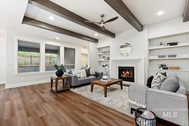 living room featuring wood finished floors, built in features, baseboards, beam ceiling, and a glass covered fireplace