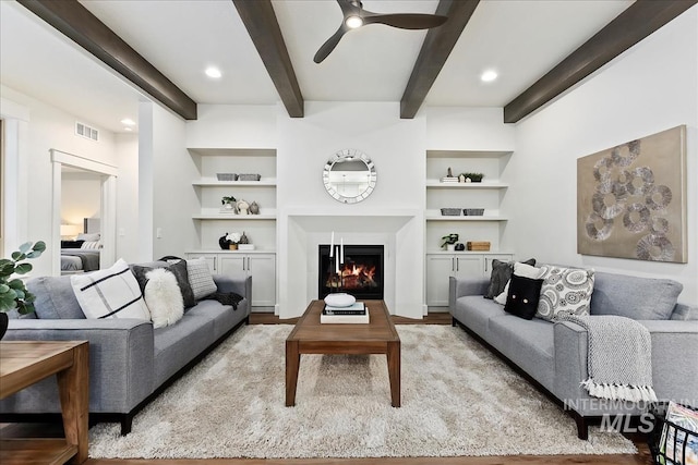 living room featuring visible vents, ceiling fan, a lit fireplace, built in shelves, and beam ceiling