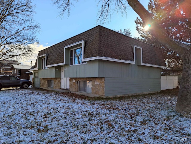 exterior space with mansard roof, a garage, a shingled roof, fence, and stone siding