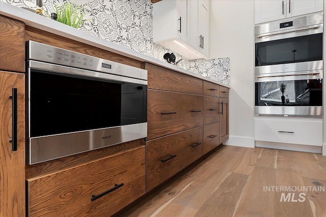 kitchen with white cabinets, stainless steel double oven, tasteful backsplash, and light hardwood / wood-style flooring