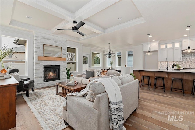 living room featuring light wood-type flooring, a brick fireplace, coffered ceiling, ceiling fan with notable chandelier, and beam ceiling