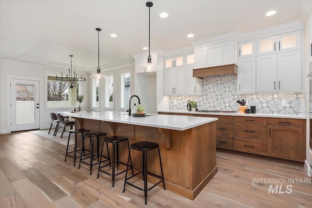 kitchen with sink, pendant lighting, a kitchen island with sink, white cabinets, and light wood-type flooring