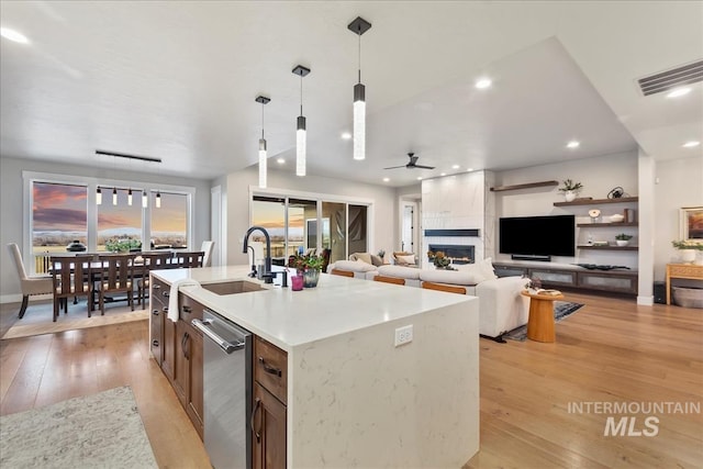 kitchen featuring a kitchen island with sink, a sink, light wood-style flooring, hanging light fixtures, and stainless steel dishwasher