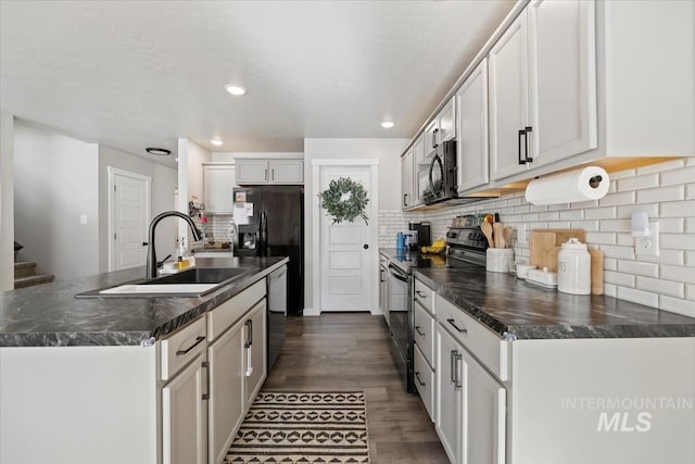 kitchen featuring dark wood-style floors, dark countertops, a sink, an island with sink, and black appliances