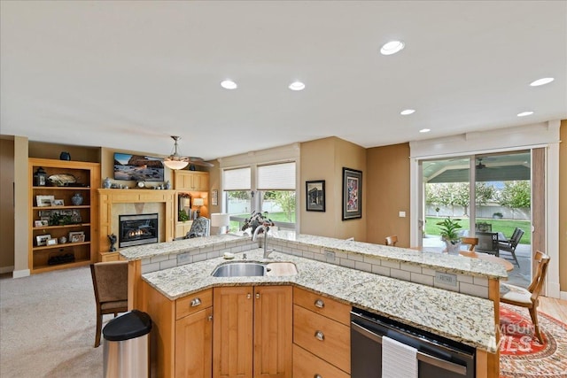 kitchen with stainless steel dishwasher, a glass covered fireplace, open floor plan, an island with sink, and a sink