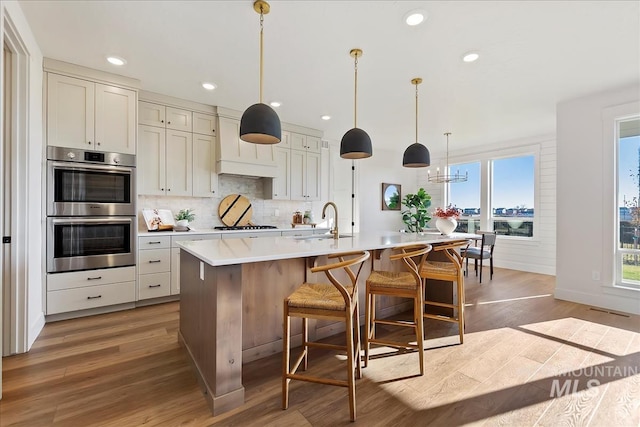 kitchen with sink, double oven, decorative light fixtures, a center island with sink, and hardwood / wood-style flooring