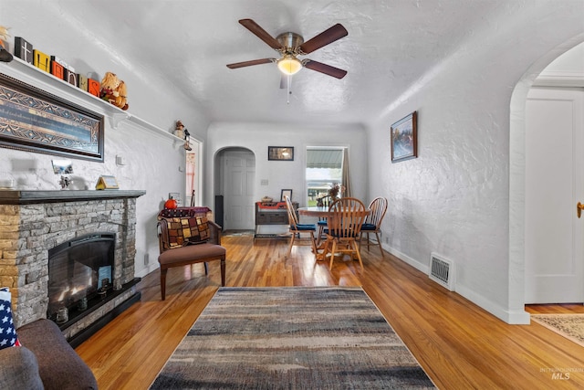 living room with a stone fireplace, a textured ceiling, light wood-type flooring, and ceiling fan