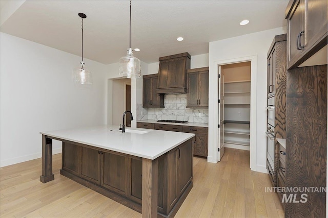 kitchen featuring sink, tasteful backsplash, a kitchen island with sink, dark brown cabinets, and light wood-type flooring