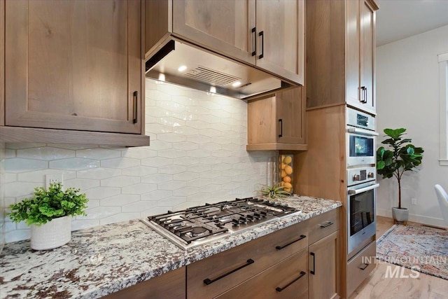 kitchen featuring under cabinet range hood, stainless steel appliances, backsplash, and light stone countertops