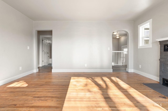 unfurnished living room featuring a brick fireplace, baseboards, arched walkways, and wood finished floors