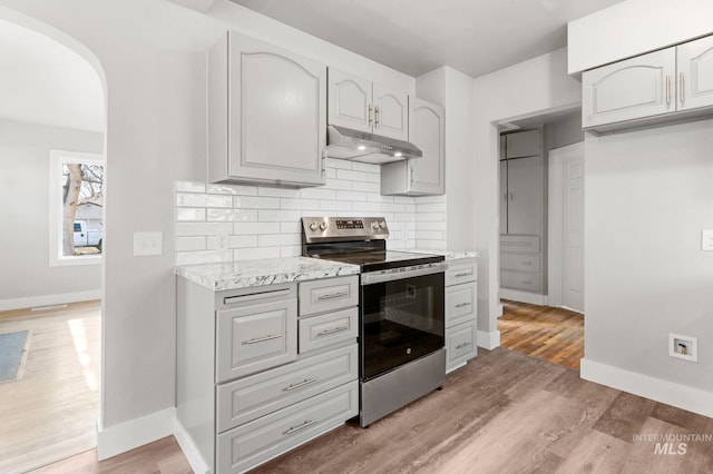 kitchen with arched walkways, backsplash, light wood-type flooring, under cabinet range hood, and stainless steel electric range