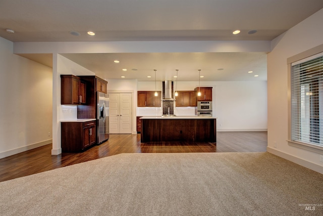 kitchen featuring wall chimney range hood, dark hardwood / wood-style floors, pendant lighting, a center island with sink, and appliances with stainless steel finishes