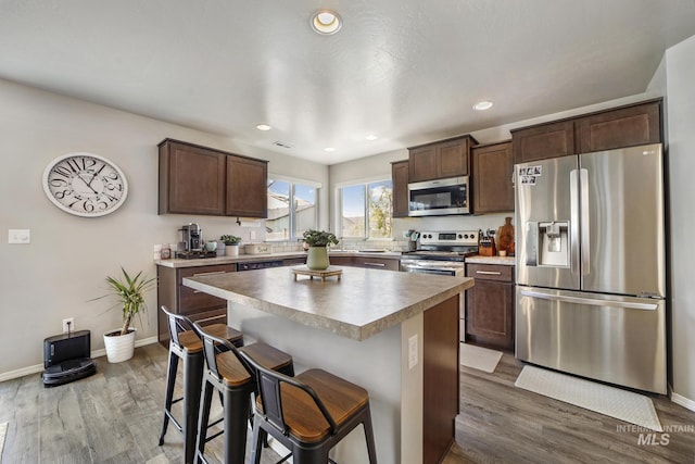 kitchen with appliances with stainless steel finishes, a breakfast bar, a kitchen island, dark brown cabinets, and hardwood / wood-style flooring