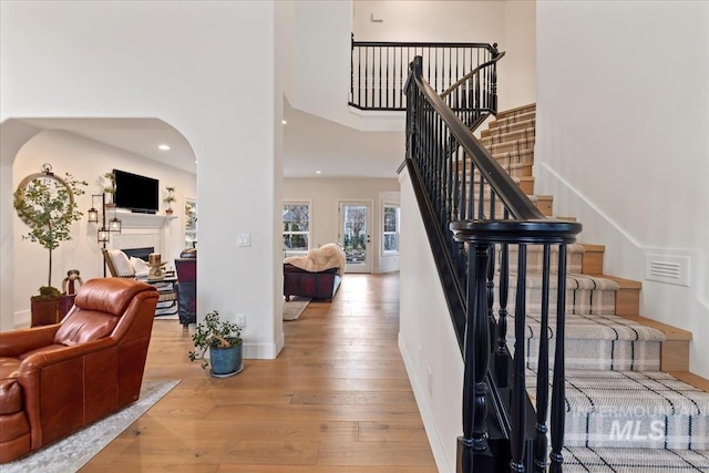 staircase featuring arched walkways, a fireplace, a towering ceiling, baseboards, and hardwood / wood-style flooring