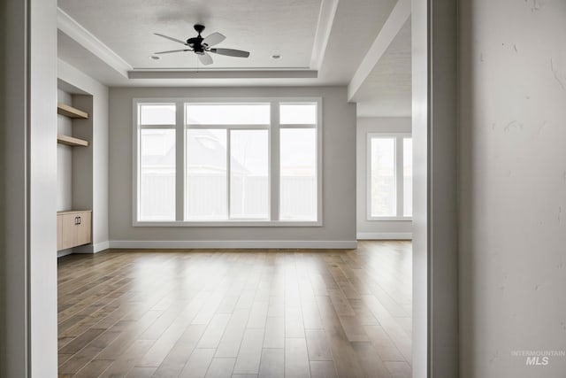 empty room with a ceiling fan, wood finished floors, built in features, baseboards, and a raised ceiling