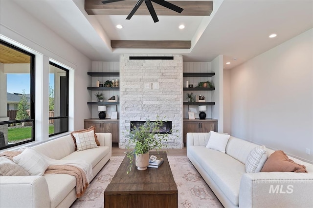 living room featuring light wood-style flooring, ceiling fan, a tray ceiling, recessed lighting, and a stone fireplace