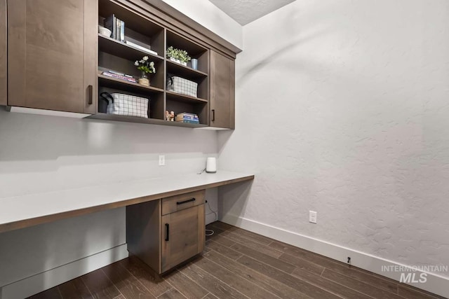 unfurnished office featuring dark wood-style floors, built in desk, a textured wall, and baseboards