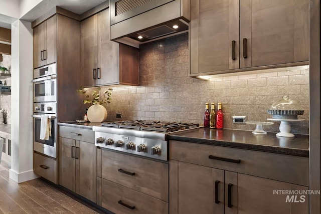 kitchen with dark wood finished floors, stainless steel appliances, custom range hood, dark countertops, and backsplash