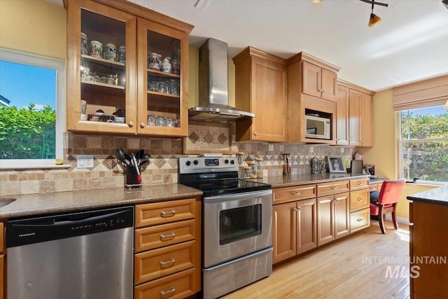 kitchen featuring wall chimney range hood, light hardwood / wood-style flooring, stainless steel appliances, dark stone counters, and tasteful backsplash