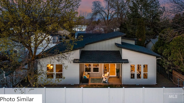 back of house at dusk featuring a fenced front yard and a shingled roof