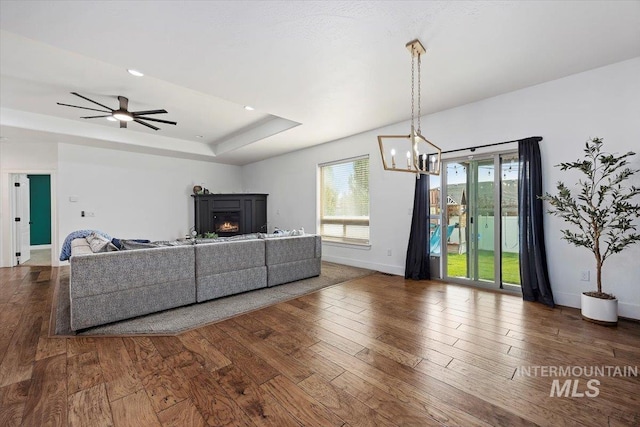 living area featuring ceiling fan with notable chandelier, wood finished floors, baseboards, a lit fireplace, and a raised ceiling