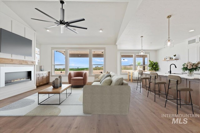 living room with light wood-style flooring, a glass covered fireplace, vaulted ceiling, and ceiling fan