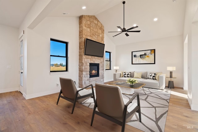 living area featuring baseboards, ceiling fan, a stone fireplace, light wood-style flooring, and high vaulted ceiling
