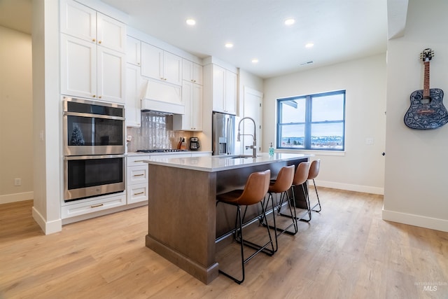 kitchen with a kitchen bar, appliances with stainless steel finishes, custom exhaust hood, white cabinets, and a kitchen island with sink