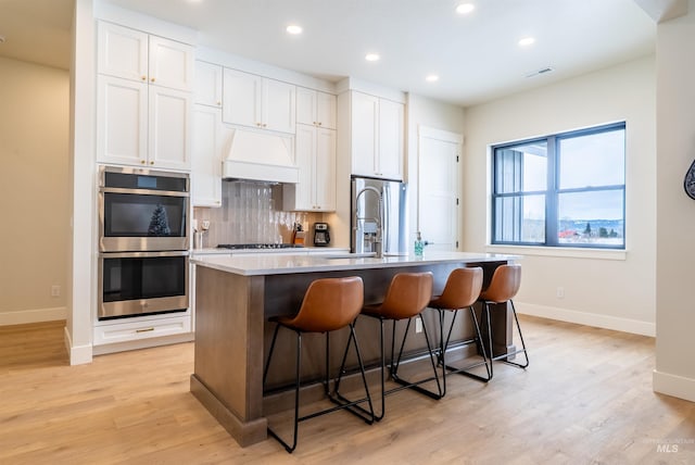kitchen with custom exhaust hood, an island with sink, a kitchen bar, double oven, and tasteful backsplash