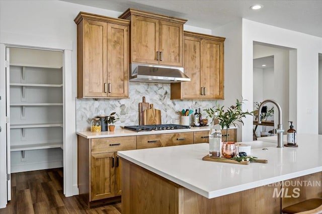 kitchen featuring stainless steel gas cooktop, brown cabinetry, under cabinet range hood, light countertops, and dark wood-type flooring