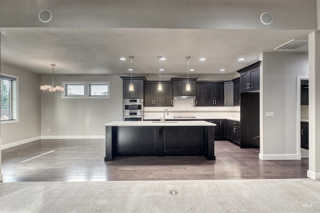 kitchen featuring tasteful backsplash, stainless steel double oven, sink, decorative light fixtures, and a center island with sink