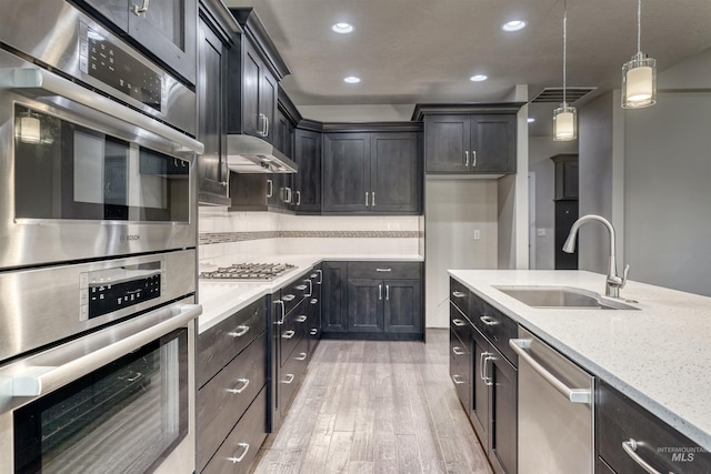 kitchen with light stone countertops, appliances with stainless steel finishes, light wood-type flooring, sink, and decorative light fixtures