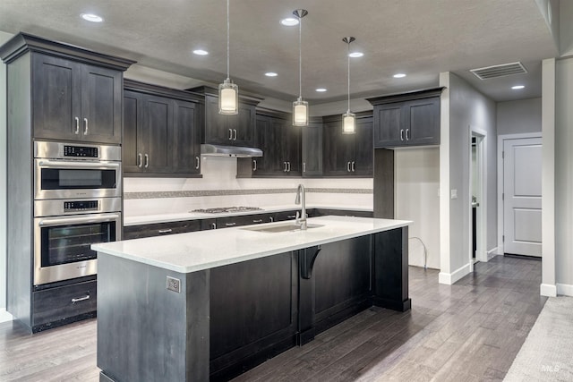 kitchen with stainless steel appliances, sink, a center island with sink, hardwood / wood-style flooring, and hanging light fixtures