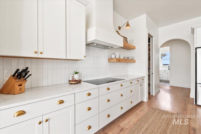 kitchen with arched walkways, black electric stovetop, custom exhaust hood, tasteful backsplash, and light wood-style floors