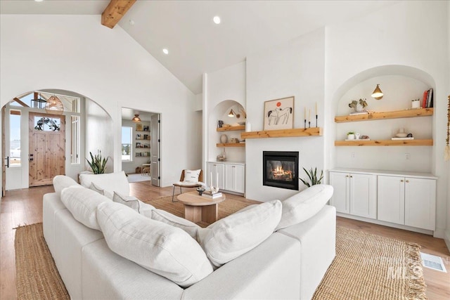 living room featuring built in shelves, beamed ceiling, a glass covered fireplace, and light wood-style floors