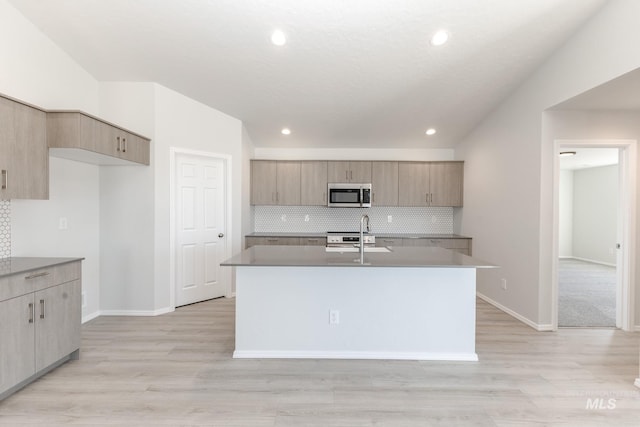 kitchen with baseboards, light wood-style flooring, an island with sink, stainless steel microwave, and tasteful backsplash