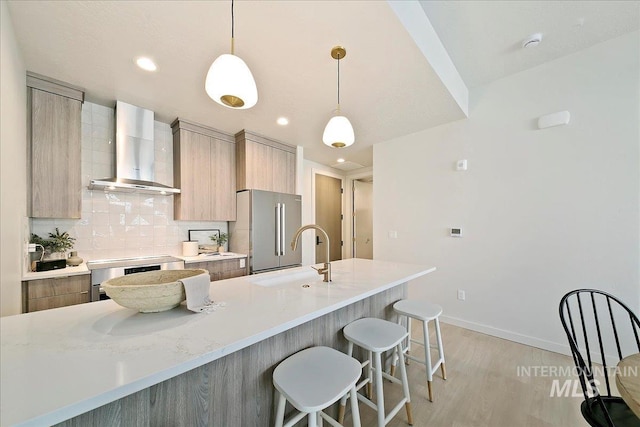 kitchen featuring high end refrigerator, backsplash, sink, wall chimney range hood, and hanging light fixtures