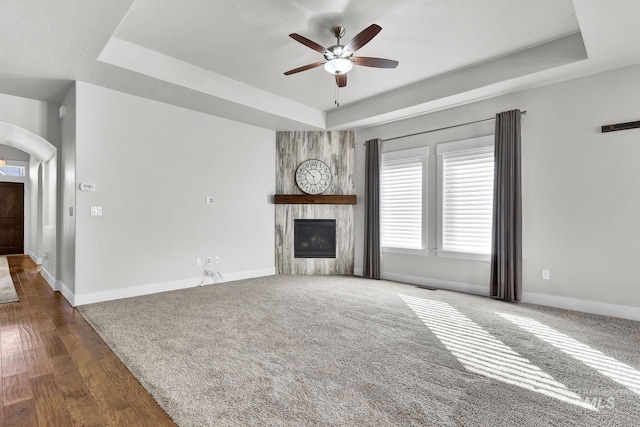 unfurnished living room with baseboards, arched walkways, a ceiling fan, a tray ceiling, and a fireplace