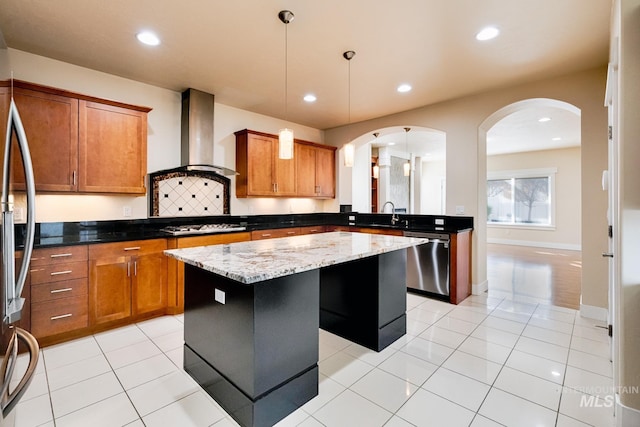 kitchen featuring brown cabinetry, arched walkways, stainless steel appliances, wall chimney exhaust hood, and a center island